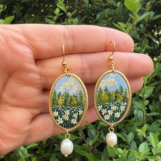 a hand holding a pair of earrings with flowers and trees painted on the side of it