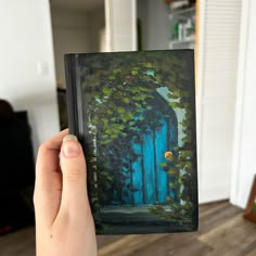 a hand holding up a small painting in front of a door with vines on it