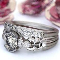 two silver rings with diamonds on them sitting in front of some flowers and fabric material