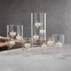 four clear glass vases filled with candles on top of a gray surface next to driftwood