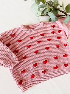 a pink sweater with red hearts on it next to some green leaves and a plant