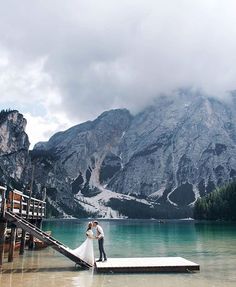 a bride and groom standing on a dock in front of a mountain lake with snow capped mountains