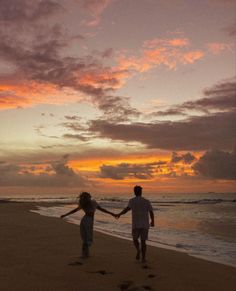 two people holding hands walking on the beach at sunset with clouds in the sky above them