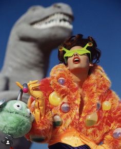 a woman in an orange fur coat and green goggles standing next to a fake dinosaur