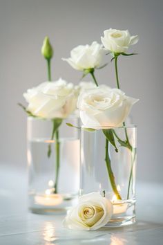 three clear vases filled with white roses on top of a marble countertop next to candles