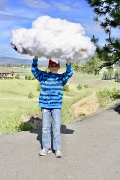 a person holding up a cloud over their head in the middle of a road with grass and trees behind them