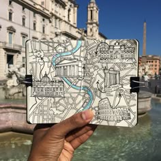 a hand holding up a small notebook with a map on it in front of a fountain