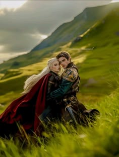 a man and woman dressed in medieval clothing hugging each other on the grass with mountains in the background