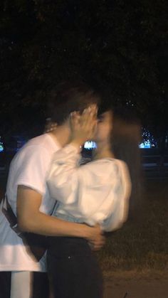 two people standing next to each other in front of trees at night, one holding the other's face