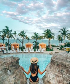 a woman in a black swimsuit and hat sitting on rocks near a pool with palm trees