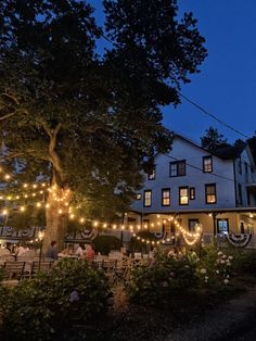 people are sitting at tables in front of a house with lights strung from the trees