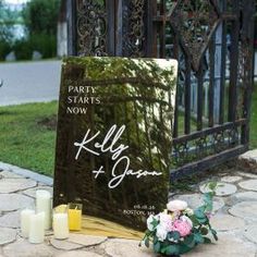 a sign with candles and flowers sitting on the ground next to some grass in front of a gate