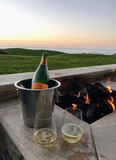 two wine glasses sitting next to an open fire pit with a bottle of champagne in it