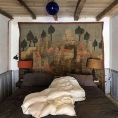 an unmade bed in front of a painting on the wall and two lamps next to it