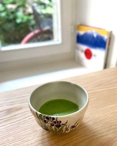 a bowl filled with green liquid sitting on top of a wooden table next to a window