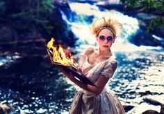 a woman holding a tray with flames in her hands near a river and waterfall wearing sunglasses