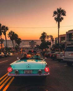 a blue car driving down a street next to the ocean at sunset with palm trees