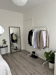 a bedroom with white walls and wooden flooring has clothes on hangers, a mirror, potted plants, and a bed