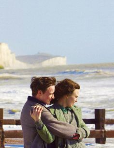 two people standing next to each other in front of the ocean with their arms around one another