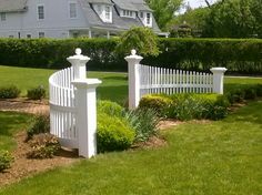 a white picket fence in front of a house with green grass and bushes around it