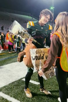 a woman holding a teddy bear on the sidelines at a football game while another person holds up a sign