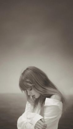 a woman standing in front of the ocean with her hair blowing in the wind and looking down