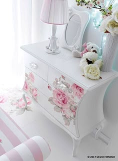 a white dresser with flowers on it next to a pink lamp and striped bedding