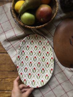 a person is holding a plate near a bowl of fruit on a table with a checkered cloth