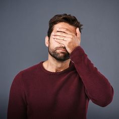 a man holding his hands to his face and covering his eyes with one hand while standing against a gray background