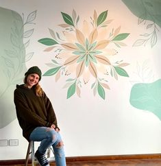 a woman sitting on a stool in front of a wall with flowers painted on it