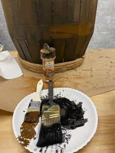 two paint brushes on a white plate next to a wooden barrel with brown substance in it