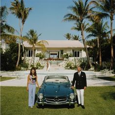 a man and woman standing next to a classic car in front of a large house