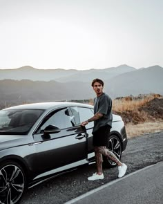 a man standing next to a parked car on the side of a road with mountains in the background