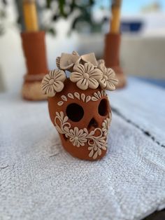 a small clay skull with flowers on its head sitting on a white towel next to two candles