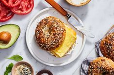 a bagel sandwich on a plate next to sliced tomatoes, avocado and bread