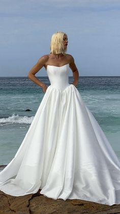 a woman in a white wedding dress standing on the rocks by the ocean with her hands on her hips