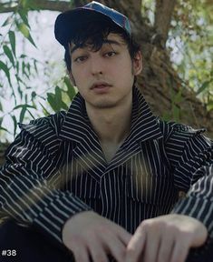 a young man sitting on the ground wearing a baseball cap and striped shirt with his hands in his pockets