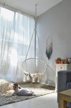 a young child laying on the floor in front of a window with curtains and a hanging chair