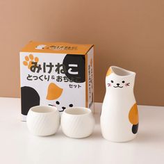 three ceramic cups and a cat figurine are on the table next to a box