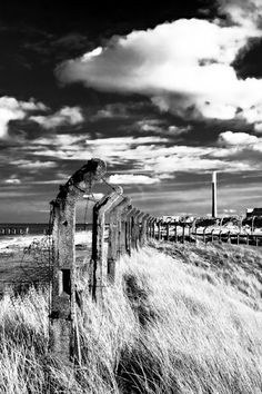 black and white photograph of an old fence on the beach with clouds in the background