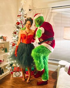 a woman standing next to a man dressed as the grinch in front of a christmas tree