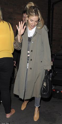 a woman in a trench coat is waving at the camera while holding her hand up