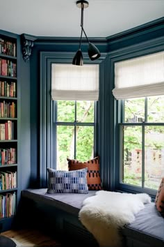 a window seat in front of two windows with bookshelves and pillows on it