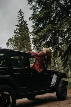 a woman leaning on the back of a black jeep parked in front of some trees