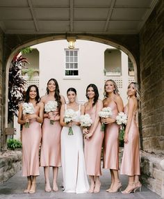 a group of women standing next to each other in front of a stone wall holding bouquets