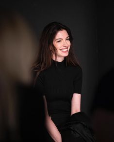 a woman in black dress sitting down and smiling at the camera with her hands on her hips