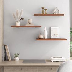 three wooden shelves on the wall above a desk with a laptop, books and vases