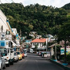 cars parked on the side of a road in front of buildings and trees with hills in the background