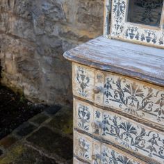 an old wooden dresser with painted drawers and mirror on it's top, in front of a stone wall