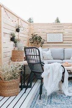 an outdoor living room with furniture and plants on the floor, in front of a wooden fence